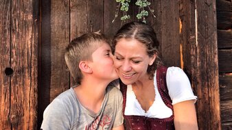 Ein Kind küsst seine Mutter in einem Trachtenkleid auf die Wange auf der Bank vor einer Almhütte | © Haus Tannenblick