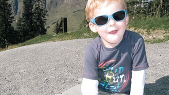 Eine Nahaufnahme eines Kindes mit Sonnenbrille auf einem Schotterweg sitzend | © Haus Tannenblick