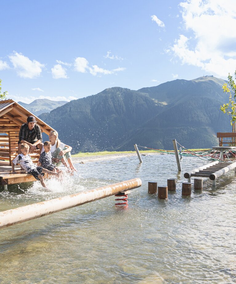 Eine Familie sitzt am Ufer eines Bergsees mit Holzhütte und planscht im Wasser in Saalbach Hinterglemm | © saalbach.com - Karin Pastererl