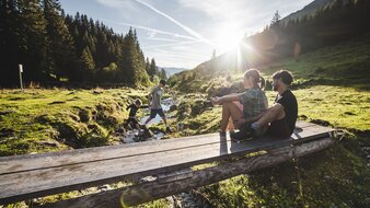 Eine wandernde Familie mit zwei Kindern rastet an einer kleinen Holzbrücke an einem Bergbach im Talschluss | © saalbach.com - Mia Knoll