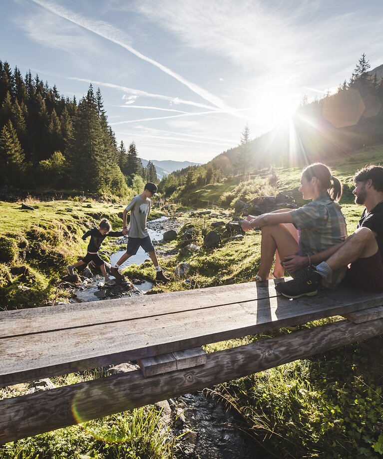 Eine wandernde Familie mit zwei Kindern rastet an einer kleinen Holzbrücke an einem Bergbach im Talschluss | © saalbach.com - Mia Knoll